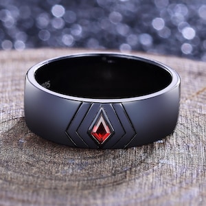 May include: A black ring with a geometric design and a red gemstone. The ring is set on a wooden surface, with a blurred background. The ring's design features a central diamond shape with a red stone.