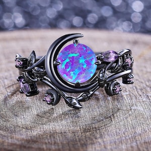 May include: A black ring with a large, round, iridescent opal stone in shades of blue and purple. The ring features a crescent moon design and is adorned with small purple gemstones and floral accents.