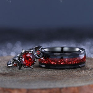 Pode incluir: Um conjunto de alianças de casamento pretas com uma gema vermelha. A aliança de noivado apresenta um design de lua crescente e a banda tem um acabamento preto escovado com detalhes vermelhos.