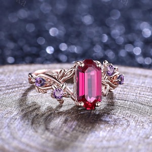 May include: A rose gold ring featuring a central emerald-cut deep red gemstone. The band is designed with intricate metal leaf motifs on both sides, accented by small, round, purple stones set within the foliage, creating a botanical aesthetic.