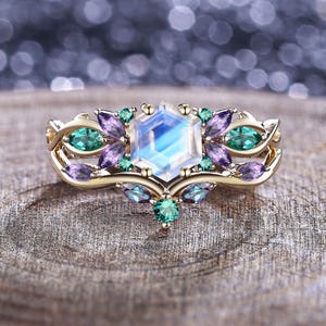 Puede incluir: Un anillo de oro con una piedra central hexagonal con un brillo iridiscente azul-blanco, rodeada de piedras preciosas verdes y moradas más pequeñas. El anillo presenta un diseño ornamental de inspiración floral.