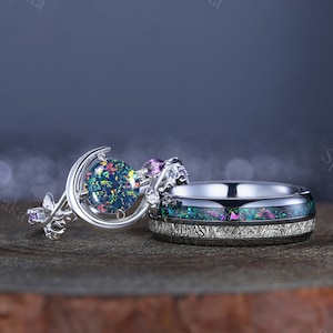 May include: A set of two silver rings with a large iridescent opal gemstone set in a crescent moon design on the engagement ring. The wedding band has a black and silver inlay with a similar iridescent opal design.