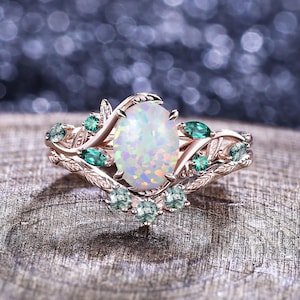 May include: Rose gold ring featuring an oval opal center stone, surrounded by green gemstones and delicate leaf accents. The ring's design incorporates a nature-inspired aesthetic, with a focus on intricate details and a vintage style.