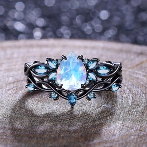 Könnte beinhalten: Ein Ring aus schwarzem Metall mit einem zentralen, birnenförmigen, irisierenden Mondstein, der von kleineren, marquise-geschliffenen blauen Steinen umgeben ist. Die Ringschiene hat ein komplexes, rankenartiges Design.