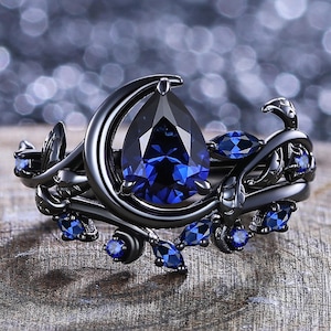 May include: Black ring with a large, pear-shaped, deep blue gemstone at the center, surrounded by smaller blue stones and a crescent moon design. The ring has a floral and vine-like pattern.