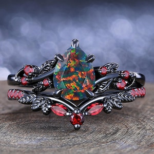 May include: Black engagement ring set with a pear-shaped opal center stone and red gemstone accents. The ring features a floral design with a black finish. The set includes a matching band with red gemstones.
