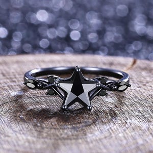 May include: A black star-shaped ring with a faceted black gemstone center. The ring has a dark metal band with decorative accents and small black gemstones. The ring is displayed on a wooden surface.