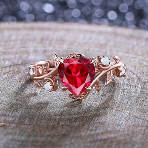 May include: Rose gold ring featuring a heart-shaped, vibrant red gemstone as the centerpiece. The ring is adorned with delicate leaf and vine detailing, and small, round, white gemstones. The ring is set on a wooden surface.