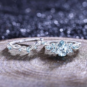 May include: A silver engagement ring set featuring a pear-shaped, light blue gemstone center stone, flanked by smaller stones. The matching band has a delicate, intertwined design with small, light blue stones. The rings are displayed on a wooden surface.