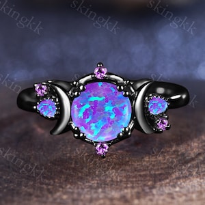 Puede incluir: Anillo negro con una gran piedra de ópalo iridiscente rodeada de pequeñas piedras moradas y tres diseños de luna creciente.