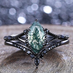Puede incluir: Un juego de anillos de compromiso negro con una piedra preciosa verde talla marquesa y piedras negras. El anillo tiene un diseño único y ornamentado con una banda a juego. El anillo está sobre una superficie de madera.