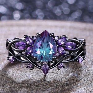 Könnte beinhalten: Ein schwarzer Ring mit einem tropfenförmigen, teal- und lilafarbenen Edelstein in der Mitte, umgeben von kleineren lila Edelsteinen. Der Ring hat ein detailliertes, blumeninspiriertes Design. Auf einer Holzoberfläche platziert.