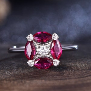 May include: A silver ring featuring a cluster of gemstones. The ring's centerpiece showcases a square arrangement of small diamonds surrounded by four oval-cut, vibrant red gemstones. Small diamonds accent the corners.