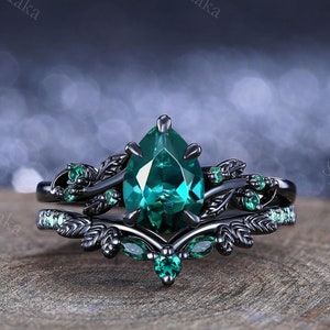 May include: A black ring set with a pear-shaped emerald green gemstone surrounded by smaller emerald green stones. The ring features a delicate leaf design.