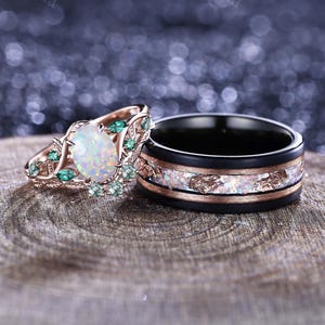 Può includere: Un anello di fidanzamento in oro rosa con una pietra centrale di opale ovale e piccole pietre preziose verdi. Accanto, un anello nero abbinato con opale e scaglie d'oro. Entrambi gli anelli sono su una superficie di legno.