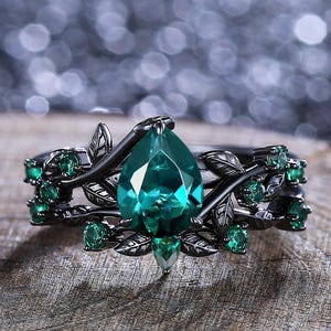 May include: A black ring with a pear-shaped emerald green gemstone surrounded by smaller emerald green gemstones and black leaves.