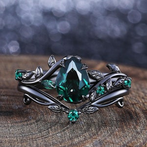 Può includere: Un set di anelli neri con una pietra preziosa verde smeraldo a forma di pera circondata da pietre preziose verde smeraldo più piccole e foglie di metallo nero.