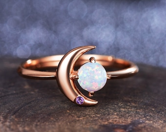 May include: A rose gold ring with a crescent moon design and a white opal gemstone. The ring has a small purple gemstone on the side.