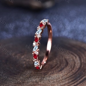 May include: Rose gold ring featuring alternating red, light blue, and white gemstones. The gemstones are marquise-cut and set in a row along the band. The ring is displayed on a wooden surface.