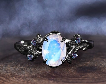 May include: A black metal ring featuring an oval, iridescent moonstone center stone. The ring has a floral design with small, purple accent stones on each side of the moonstone. The ring is set against a blurred background.