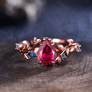 May include: A rose gold ring with a pear-shaped ruby gemstone surrounded by a delicate vine design with smaller blue gemstones.