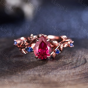 Könnte beinhalten: Ein Verlobungsring aus Roségold mit einem birnenförmigen Rubin-Edelstein und zwei kleineren blauen Saphiren, die in einem zarten Blattdesign gefasst sind.