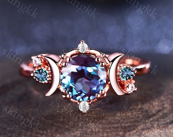May include: Rose gold ring featuring a large, round, color-changing alexandrite gemstone. The ring has a celestial design with crescent moon accents and small accent stones. The ring's band is a warm rose gold color, and the gemstones are a mix of blue and white.