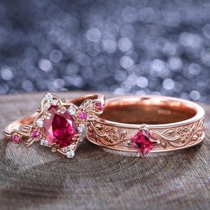 May include: Two rose gold rings. One ring features a pear-shaped, red gemstone surrounded by smaller gems. The other ring has a square red gemstone and an engraved floral design. The rings are displayed on a wooden surface.