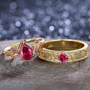 以下が含まれることがあります： 2つのゴールドリング。1つのリングは洋梨型の赤い宝石と花のアクセントが特徴です。もう1つのリングは正方形の赤い宝石と葉の模様があります。どちらのリングも木製の表面にあります。