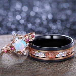May include: A pair of wedding bands. The engagement ring features a pear-shaped opal gemstone surrounded by smaller diamonds and pink gemstones. The wedding band is black with a copper-colored inlay.