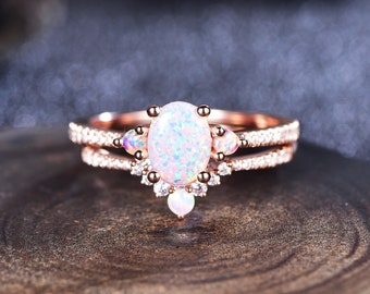 May include: A rose gold ring set with a large oval-shaped opal gemstone. The opal is surrounded by smaller clear gemstones and the band is accented with a row of smaller gemstones.