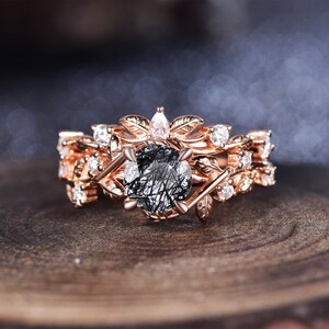 May include: A rose gold engagement ring with a black and white gemstone surrounded by smaller clear gemstones. The ring features a delicate leaf design.