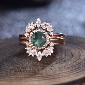 May include: A rose gold ring set with a green gemstone surrounded by a halo of smaller diamonds. The ring is accented with a delicate floral design of smaller diamonds.