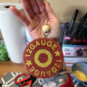 May include: A round, red air freshener with the text "12 GAUGE" in gold lettering. The air freshener has a gold circle in the center and gold star accents. It hangs from a leather cord with a wooden bead.