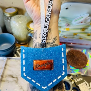 Puede incluir: Un ambientador de fieltro azul en forma de bolsillo con costuras blancas y un parche Wrangler de cuero marrón. Cuelga de un anillo negro y una cinta blanca y negra con estampado de vaca. El fondo está borroso.