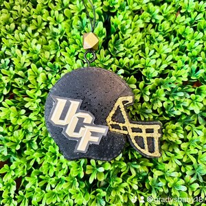 May include: A black and gold football helmet ornament with the letters "UCF" in gold on the side. The ornament is hanging from a black cord with a gold bead at the top.