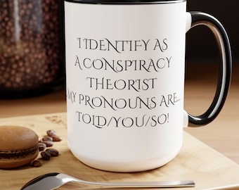 May include: White ceramic coffee mug with a black rim and handle. The mug has black text that reads "I identify as a conspiracy theorist. My pronouns are... told you so!"