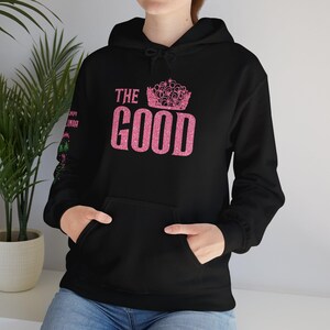 Pode incluir: Hoodie preto com um design de glitter rosa que diz "THE GOOD" com uma coroa acima do texto.