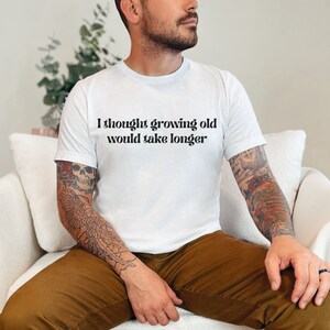 Puede incluir: Una camiseta blanca con el texto "I thought growing old would take longer" impreso en la parte delantera.