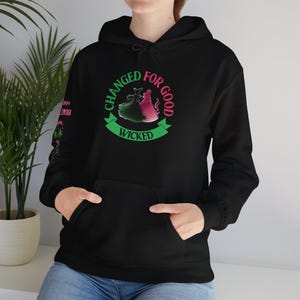 Pode incluir: Camisola de moletom com capuz preta com um design gráfico verde e rosa que diz "Changed for Good Wicked".