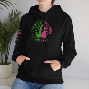 Pode incluir: Hoodie preto com uma estampa gráfica verde e rosa que diz "Changed for Good Wicked". O gráfico apresenta duas mulheres em silhueta com um relógio atrás delas.