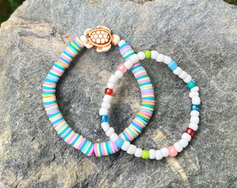 May include: Two colourful beaded bracelets with a turtle charm. The bracelet on the left is made of flat, colourful beads and the bracelet on the right is made of small, white beads with accents of red, green, and blue.