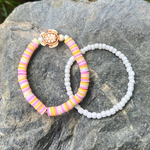 May include: Two bracelets, one white and one with pink, yellow, and white beads. The beaded bracelet has a small white turtle charm.