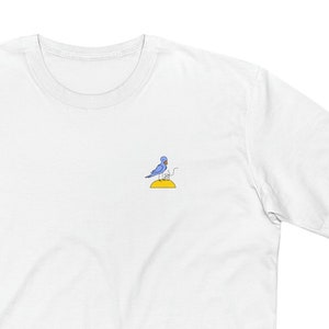 May include: White t-shirt with a blue bird embroidered on the front. The bird is standing on a yellow circle.