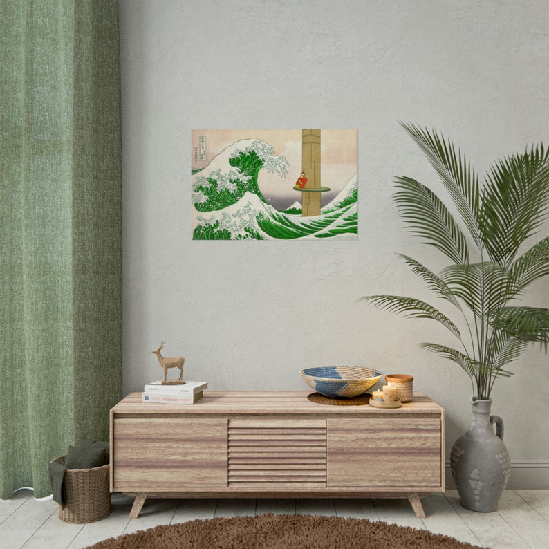 May include: A wood framed print of a large wave with a small figure standing on a platform in the distance. The print is hanging on a light beige wall above a wooden cabinet with a deer figurine and books on top.