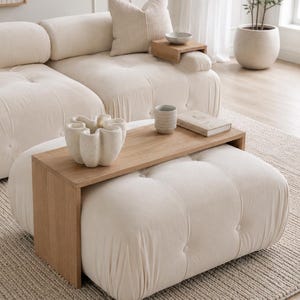 May include: A light oak wood sofa arm tray table with a rectangular top and two side panels, positioned over a tufted cream-coloured ottoman. The tray holds a white sculptural ceramic vase, a textured ceramic mug, and a small closed book.