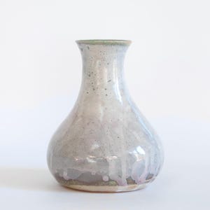 Puede incluir: Un jarrón de cerámica hecho a mano con cuerpo en forma de pera y cuello estrecho. El jarrón presenta un acabado vidriado moteado en tonos grises, con sutiles toques de púrpura y un borde verde. La superficie brillante refleja la luz.