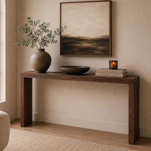 May include: A dark wood console table featuring a rectangular top and two side legs. On the table sits a round ceramic vase with green branches, a dark shallow bowl, two stacked books, and a glass candle holder. A square framed abstract painting hangs above.