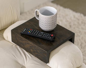 May include: A dark brown wooden armrest tray with a white ceramic mug and a black remote control on top. The tray is attached to a white couch.