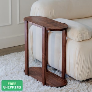 Puede incluir: Una mesa auxiliar ovalada de color marrón oscuro con un diseño minimalista. La mesa tiene una base y una parte superior rectangulares, conectadas por dos soportes verticales. La mesa es de madera y está colocada junto a un sofá crema.